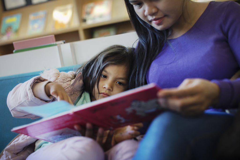 En kvinna med lila tröja som läser en bilderbok tillsammans med ett barn i femårsåldern. Båda ser allvarliga ut. Barnet pekar på en sida i boken.