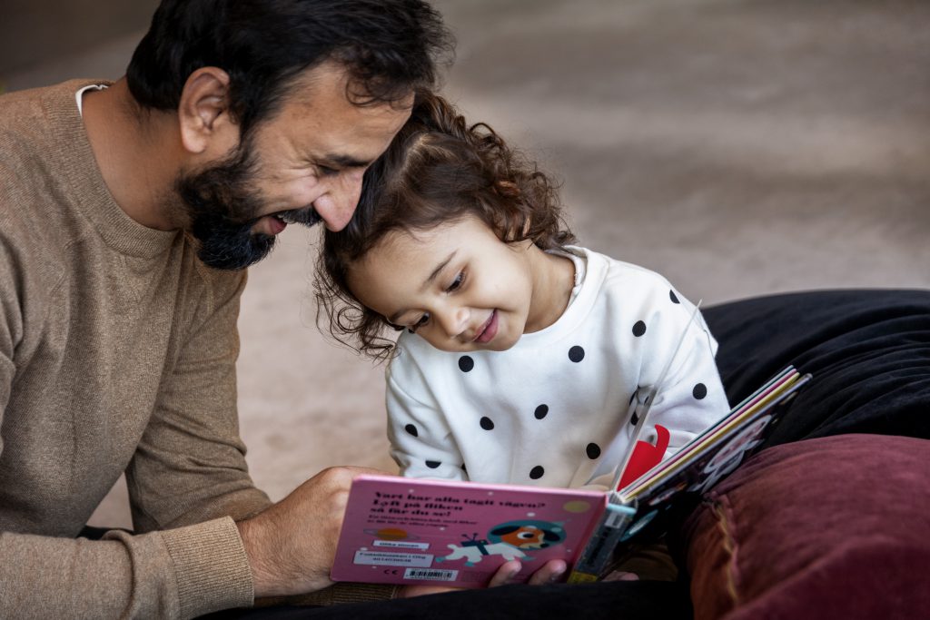 En man med mörkt skägg som tittar i en bilderbok tillsammans med ett barn i treårsåldern. Båda ser glada ut.