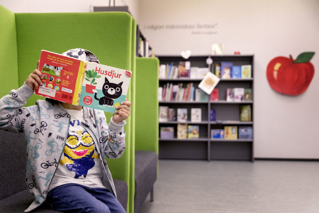 Ett barn som håller upp en pekbok framför sitt ansikte. Barnet är på biblioteket på avdelningen för tillgänglig litteratur, den så kallade äppelhyllan. På bokens framsida syns en bild av en katt.